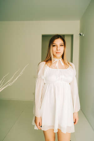 Portrait of attractive caucasian young woman in a white dress standing in an empty room with white walls.の写真素材