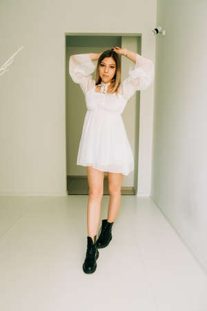 Portrait of attractive caucasian young woman in a white dress standing in an empty room with white walls.の写真素材
