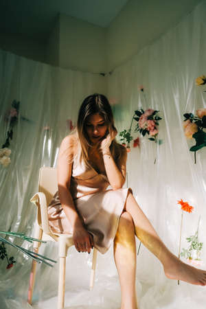 Portrait of attractive caucasian woman in silky dress, sitting in a sunny room with flowers on a wall.の写真素材