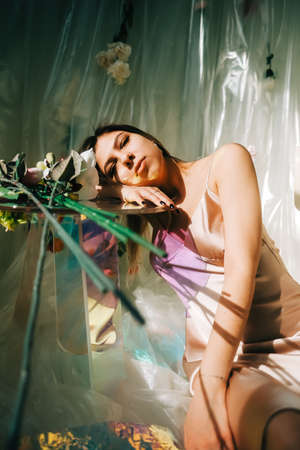 Portrait of attractive caucasian woman in silky dress, sitting near table in a sunny room with flowers on a wall.の写真素材