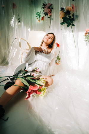 Portrait of attractive caucasian woman in short white dress, sitting on a floor in a sunny room with flowers on a wall.の写真素材