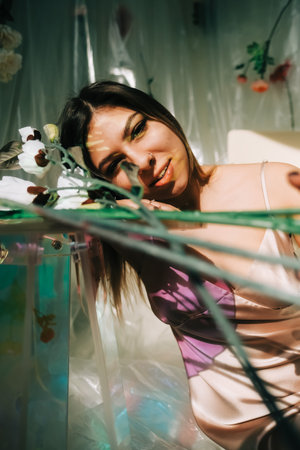 Portrait of attractive caucasian woman in silky dress, sitting near table in a sunny room with flowers on a wall.の写真素材