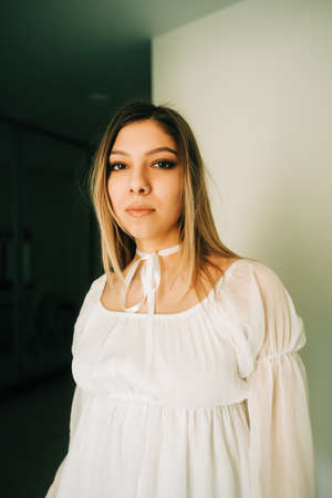 Portrait of attractive caucasian young woman in a white dress standing in an empty room with white walls.の写真素材