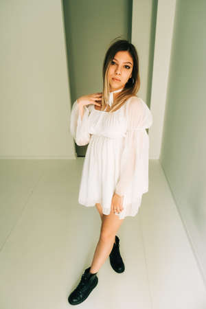 Portrait of attractive caucasian young woman in a white dress standing in an empty room with white walls.の写真素材