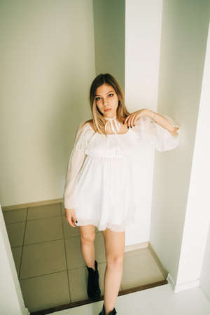 Portrait of attractive caucasian young woman in a white dress standing in an empty room with white walls.の写真素材