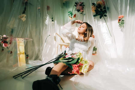Portrait of attractive caucasian woman in short white dress, sitting on a floor in a sunny room with flowers on a wall.の写真素材