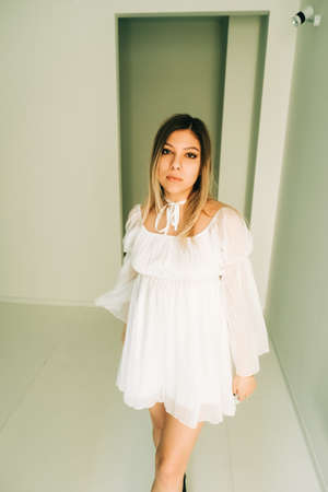 Portrait of attractive caucasian young woman in a white dress standing in an empty room with white walls.の写真素材