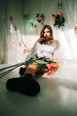 Portrait of attractive caucasian woman in short white dress, sitting on a floor in a sunny room with flowers on a wall.の写真素材