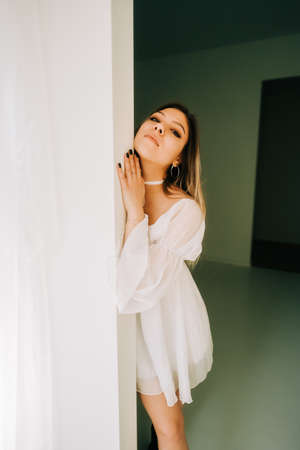 Portrait of attractive caucasian young woman in a white dress standing in an empty room with white walls.の写真素材
