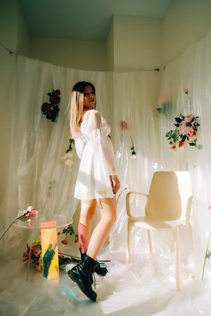 Portrait of attractive caucasian woman in short white dress, posing in a sunny room with flowers on a wall.の写真素材