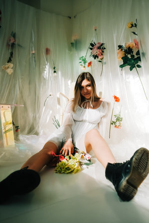 Portrait of attractive caucasian woman in short white dress, sitting on a floor in a sunny room with flowers on a wall.の写真素材