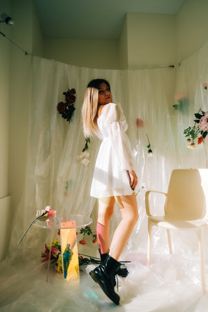 Portrait of attractive caucasian woman in short white dress, posing in a sunny room with flowers on a wall.の写真素材
