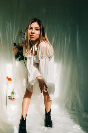 Portrait of attractive caucasian woman in short white dress, posing in a sunny room with flowers on a wall.の写真素材
