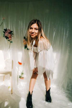 Portrait of attractive caucasian woman in short white dress, posing in a sunny room with flowers on a wall.の写真素材