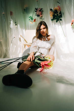 Portrait of attractive caucasian woman in short white dress, sitting on a floor in a sunny room with flowers on a wall.の写真素材