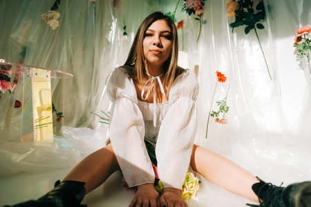 Portrait of attractive caucasian woman in short white dress, sitting on a floor in a sunny room with flowers on a wall.の写真素材