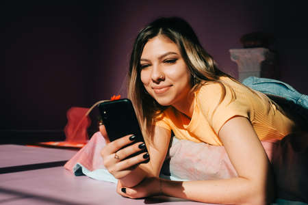 Smiling young attractive caucasian woman using mobile phone in cozy bedroom in sunlight.の写真素材