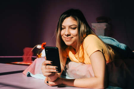 Smiling young attractive caucasian woman using mobile phone in cozy bedroom in sunlight.の写真素材