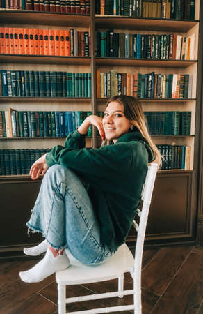 Young caucasian cheerful woman student sitting in a white chair near bookshelf in library.の写真素材
