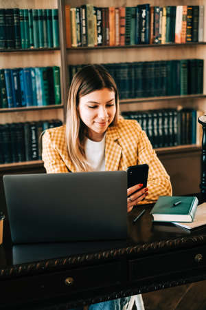 Portrait of young woman college student using mobile phone, preparing for test exam, writing essay...