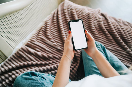 Woman holding mobile phone with white screen mock up, resting on a sofa in living room at home.の写真素材