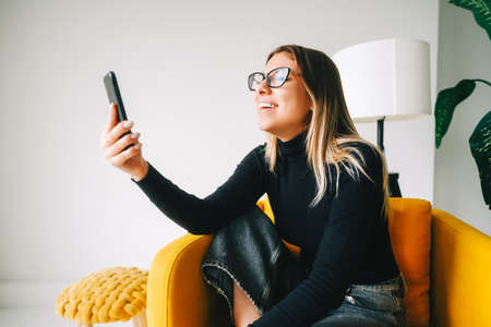 Young cheerful attractive woman in eyeglasses using mobile phone at home.の写真素材