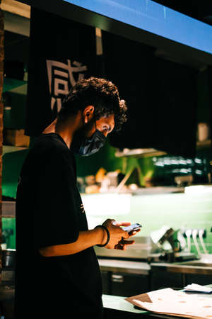 Young caucasian man asian bistro worker using mobile phone accepts online order for delivery or checks the order of guests in the hall.の写真素材