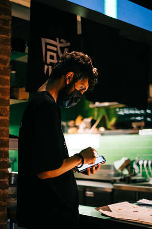 Young caucasian man asian bistro worker using mobile phone accepts online order for delivery or checks the order of guests in the hall.の写真素材