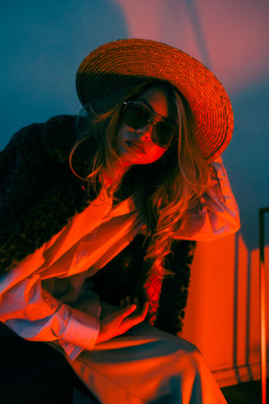 Portrait of attractive caucasian woman in eyeglasses posing in armchair wearing stylish outfit in orange light.の写真素材