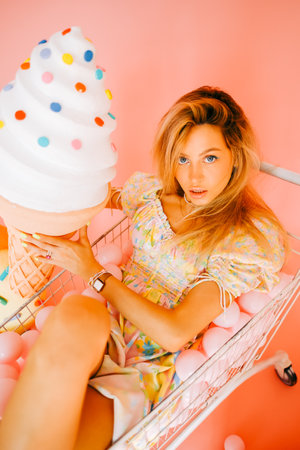 Cheerful attractive caucasian woman fashion model with blond hair in shopping cart eating big ice cream, on pink background with figures of sweets and candies.の写真素材