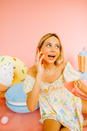 Portrait of attractive caucasian woman with blond hair talking using mobile phone, on pink background with figures of sweets and candies.の写真素材