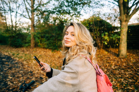 Young attractive caucasian woman using mobile phone, ride on bicycle in the park on warm day in the fall.の写真素材