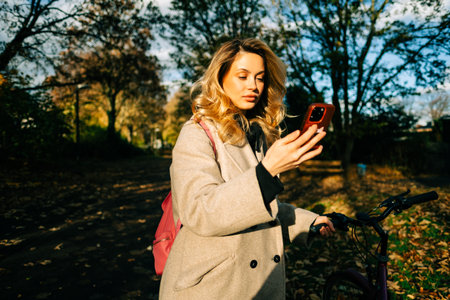 Young attractive caucasian woman using mobile phone, ride on bicycle in the park on warm day in the...