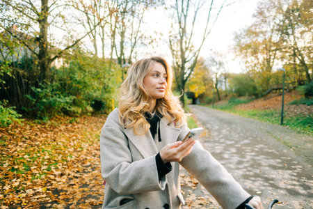 Young attractive caucasian woman using mobile phone, ride on bicycle in the park on warm day in the fall.の写真素材