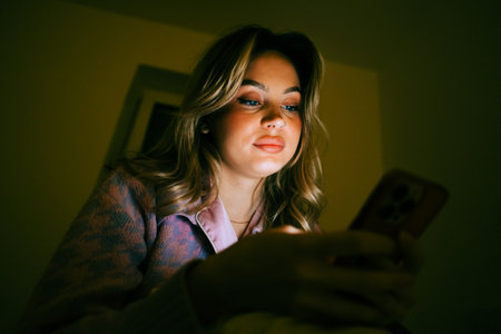 Young caucasian woman using smartphone, sitting on a bed at home in the evening.の写真素材