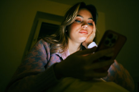 Young caucasian woman using smartphone, sitting on a bed at home in the evening.の写真素材