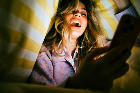 Young cheerful caucasian woman using smartphone, lies under a blanket on a bed at home in the evening.の写真素材