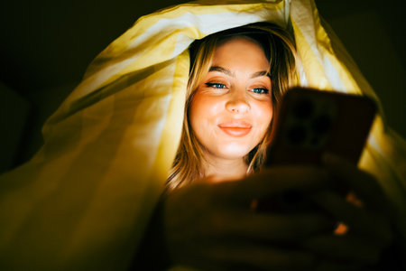 Young smiling woman using smartphone, lies under a blanket on a bed at home in the evening.の写真素材