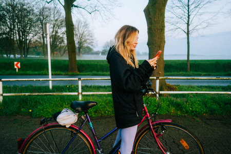 Young attractive caucasian woman using mobile phone, ride on bicycle in the park in sunset with fog .の写真素材