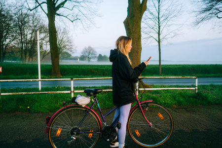 Young attractive caucasian woman using mobile phone, ride on bicycle in the park in sunset with fog .の写真素材
