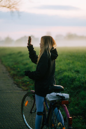 Young attractive caucasian woman making photo of beautiful view of sunset with fog while ride on bicycle in the park .の写真素材