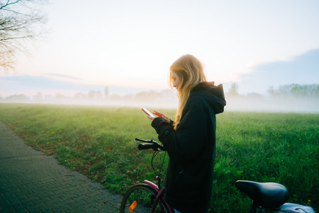 Young attractive caucasian woman using mobile phone, ride on bicycle in the park in sunset with fog .の写真素材
