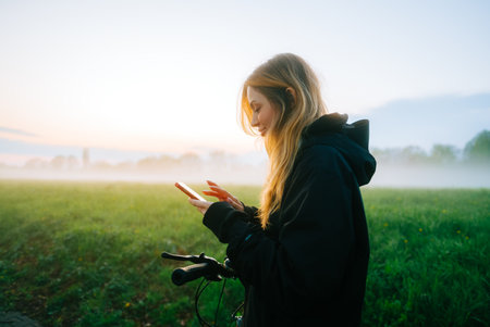 Young attractive caucasian woman using mobile phone, ride on bicycle in the park in sunset with fog .の写真素材