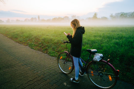 Young attractive caucasian woman using mobile phone, ride on bicycle in the park in sunset with fog .の写真素材