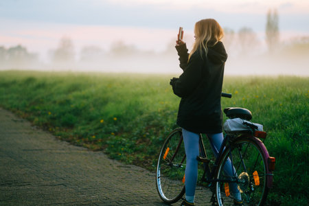 Young attractive caucasian woman making photo of beautiful view of sunset with fog while ride on bicycle in the park .の写真素材