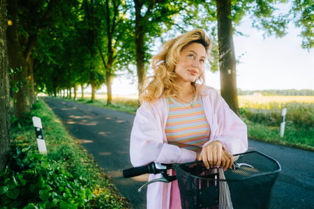 Portrait of young caucasian woman with blond hair riding on bicycle among agricultural fields in countryside in summer day.の写真素材