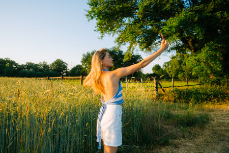 Young attractive cheerful woman making selfie photo using mobile phone at the countryside field of wheat in summer day at sunset.の写真素材
