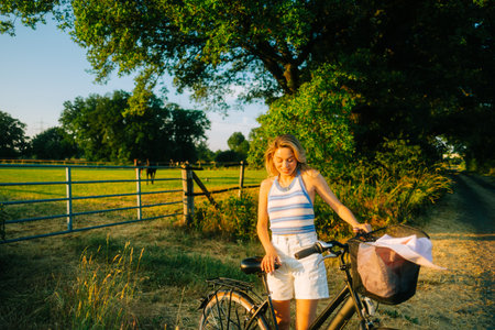 Young attractive caucasian woman with bicycle near horses pasture in summer day at sunset.の写真素材