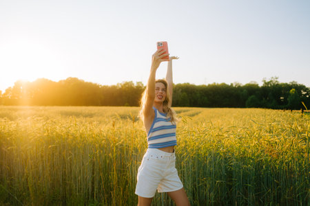 Young attractive cheerful woman making selfie photo using mobile phone at the countryside field of wheat in summer day at sunset.の写真素材