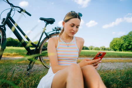 Young caucasian woman using mobile phone outdoor, sitting on grass at the countryside field, reading book or using social media.の写真素材